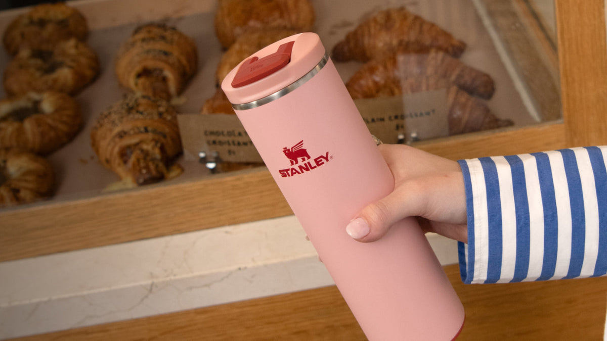 Person holding a pink travel mug with a brand logo in front of pastries.